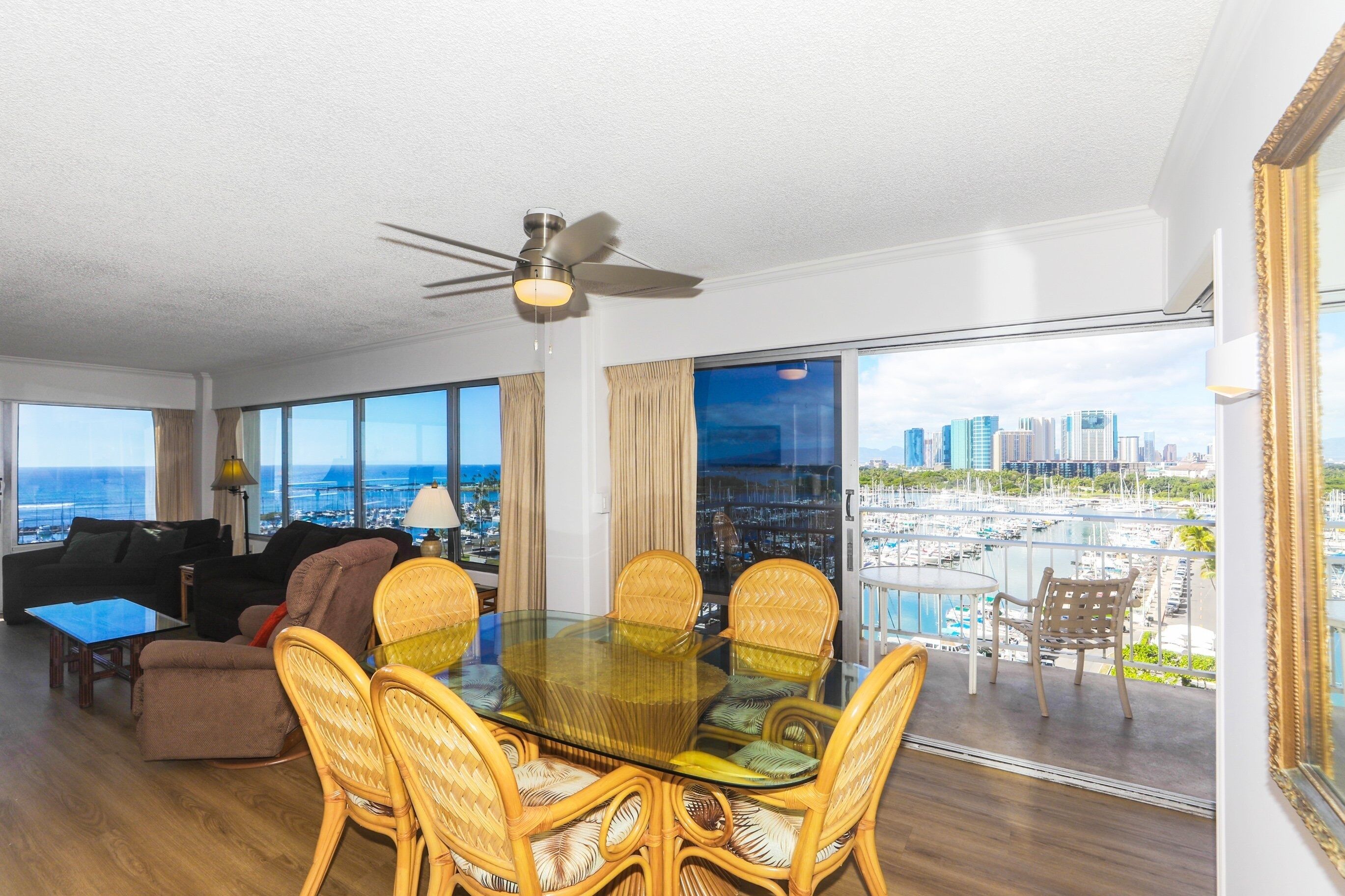 A spacious living room with a glass dining table surrounded by wicker chairs, a ceiling fan, and large windows offering a city view.