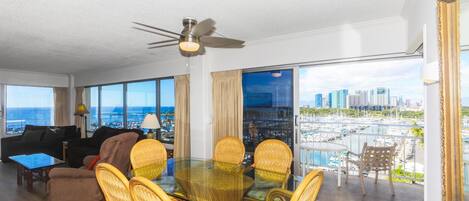 A spacious living room with a glass dining table surrounded by wicker chairs, a ceiling fan, and large windows offering a city view.