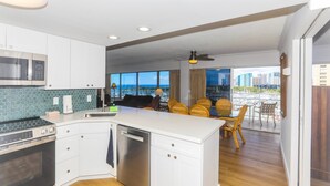 A modern kitchen with white cabinets, a stainless steel microwave, and a dishwasher. There is a dining area with wicker chairs and a view of the city through large windows. - Ilikai Tower 943 Condo - Walk to the Beach, Shops & Restaurants! by RedAwning (Honolulu)