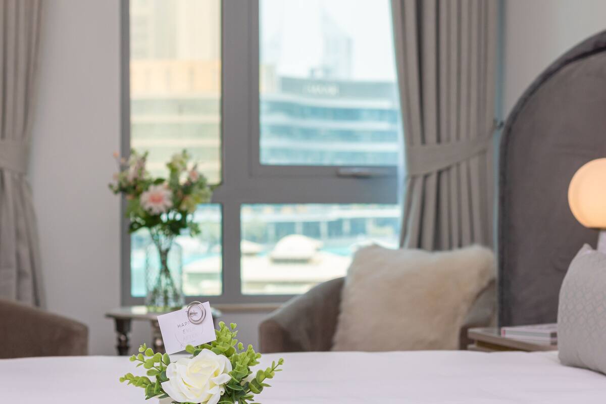 A hotel room with a bed, a nightstand, a lamp, a window with a view of a building, and a vase with flowers.