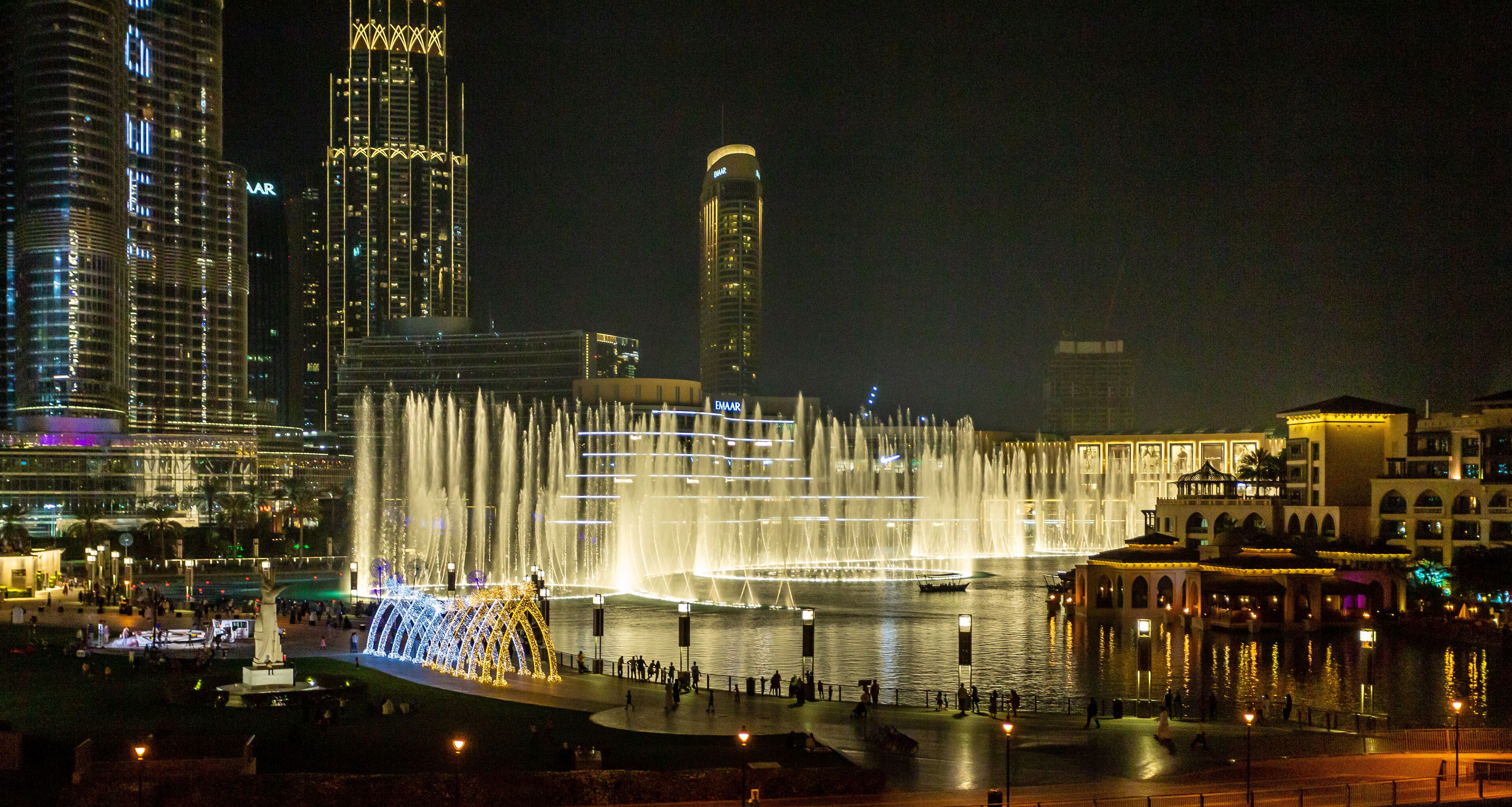 A nighttime cityscape with a prominent water fountain display, high-rise buildings, and a well-lit waterfront area.