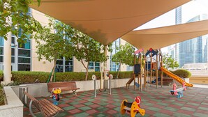 Un terrain de jeux avec un banc en bois, une balançoire et un toboggan, entouré d’un bâtiment doté de grandes fenêtres et dont l’arrière-plan est constitué d’une vue sur la ville.