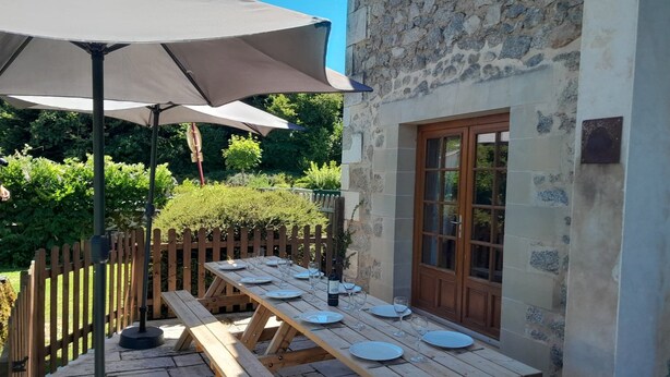 Zona pranzo all'aperto con tavoli e panche in legno, un edificio in pietra con una porta in legno e un ombrellone che offre ombra.