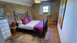 Una camera da letto con una parete in pietra, un letto con struttura in metallo, un comò, una finestra con tende e uno sgabello.