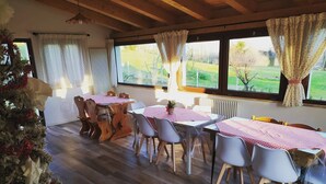 Un espace repas avec des tables et des chaises en bois, de grandes fenêtres avec des rideaux et une vue sur un paysage verdoyant.