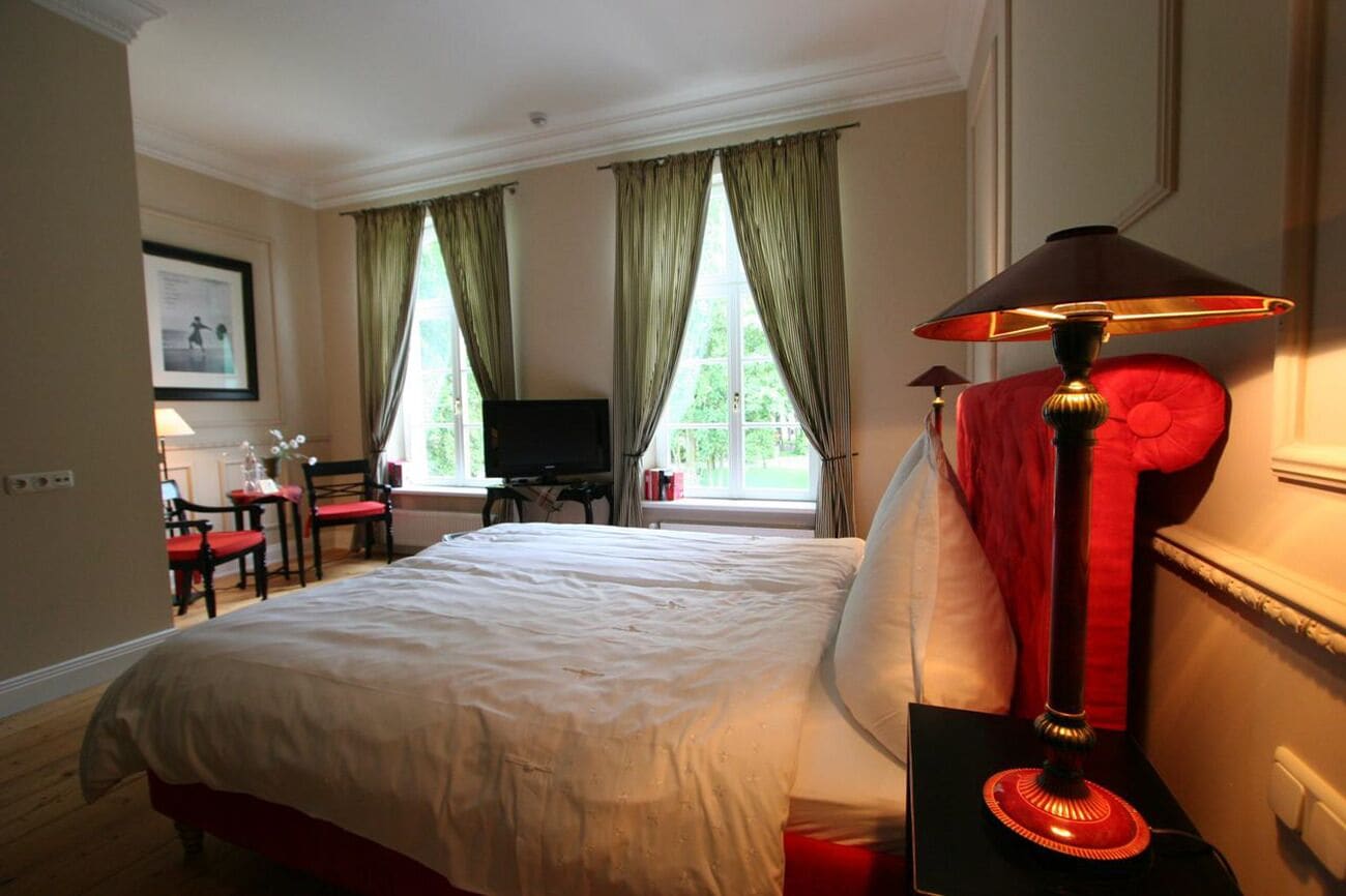 A hotel room with a large bed, two windows with green curtains, a desk with a chair, and a red lamp.