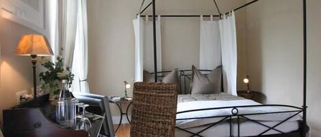A bedroom with a four-poster bed, a wicker chair, a desk with a lamp, and a framed picture on the wall.