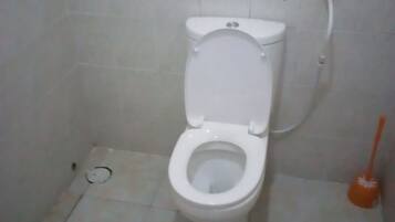 A standard white toilet in a small, tiled bathroom with a visible drain and a plunger on the floor.