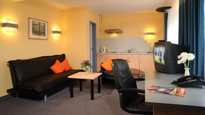 A hotel room with a black leather sofa, a small wooden table, a black leather chair, a desk with a telephone, and a television. - Apartmenthaus Triton (Cologne)