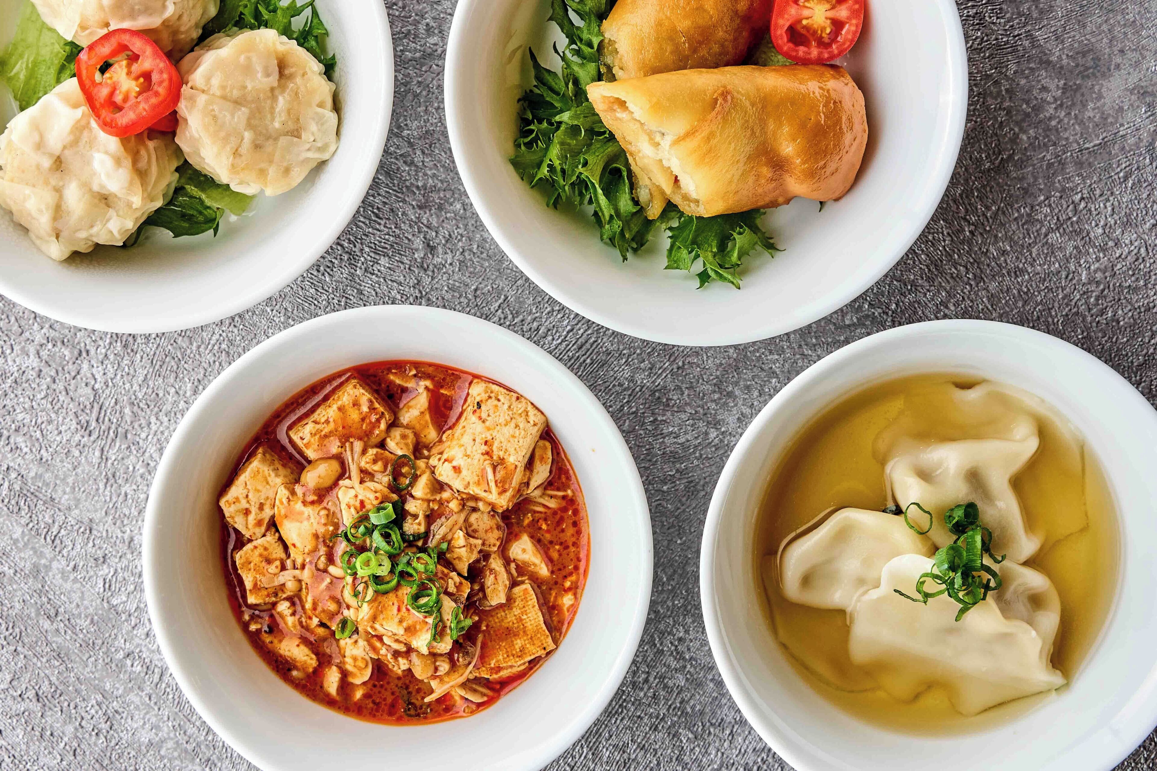 A table with four dishes: dumplings, a salad with a bread roll, a spicy tofu dish, and a soup with dumplings.