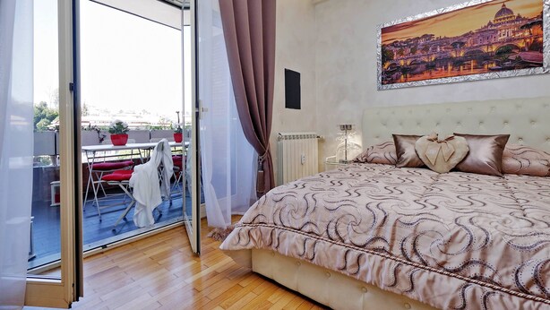 A bedroom with a large bed, a painting of a cityscape, and a view through a glass door to an outdoor area with a table and chairs. - Luxury Rome Guest House (Rome)