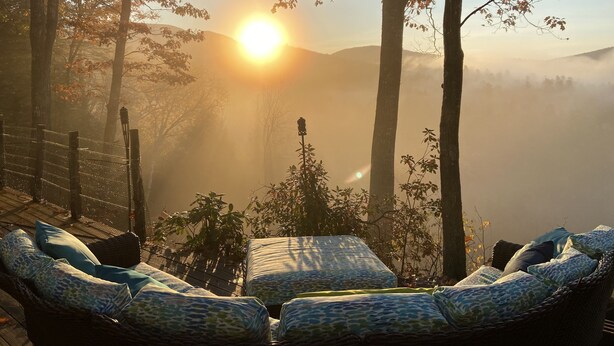 A wicker lounge chair with patterned cushions, a wooden fence, and a view of a forest with mist-covered hills and a setting sun.