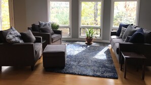 A living room with two sofas, a coffee table, and a potted plant. There are three large windows providing natural light.