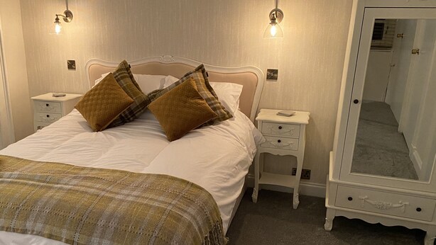 A neatly made bed with two pillows, a bedside table with a lamp, and a full-length mirror. - The Covenanter Hotel (Cupar)