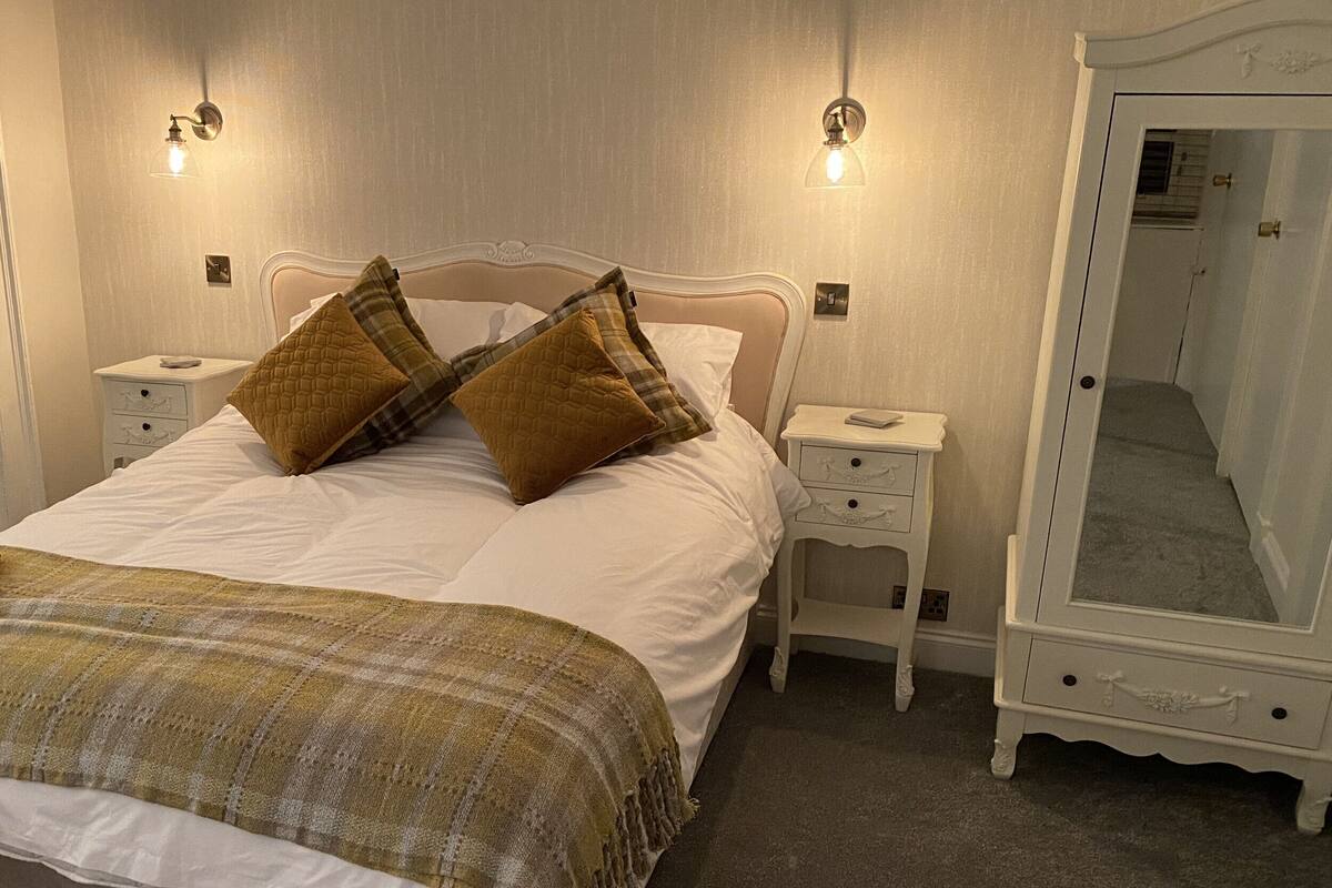 A neatly made bed with two pillows, a bedside table with a lamp, and a full-length mirror.