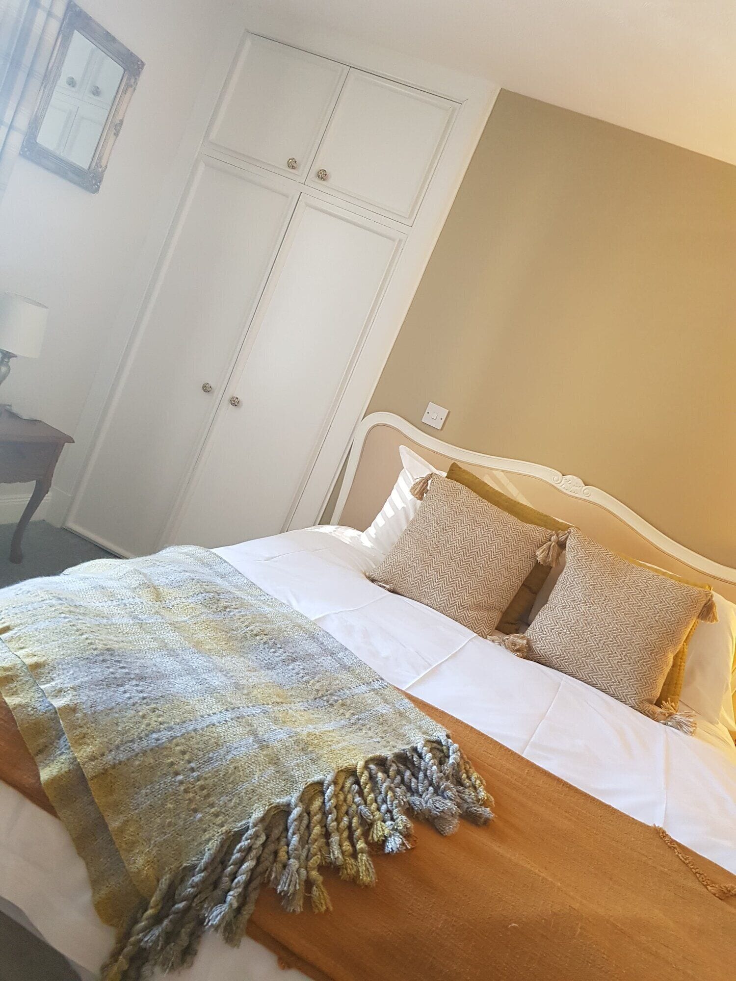 A neatly made bed with a patterned throw blanket and pillows, a white wardrobe, and a wall-mounted light switch.