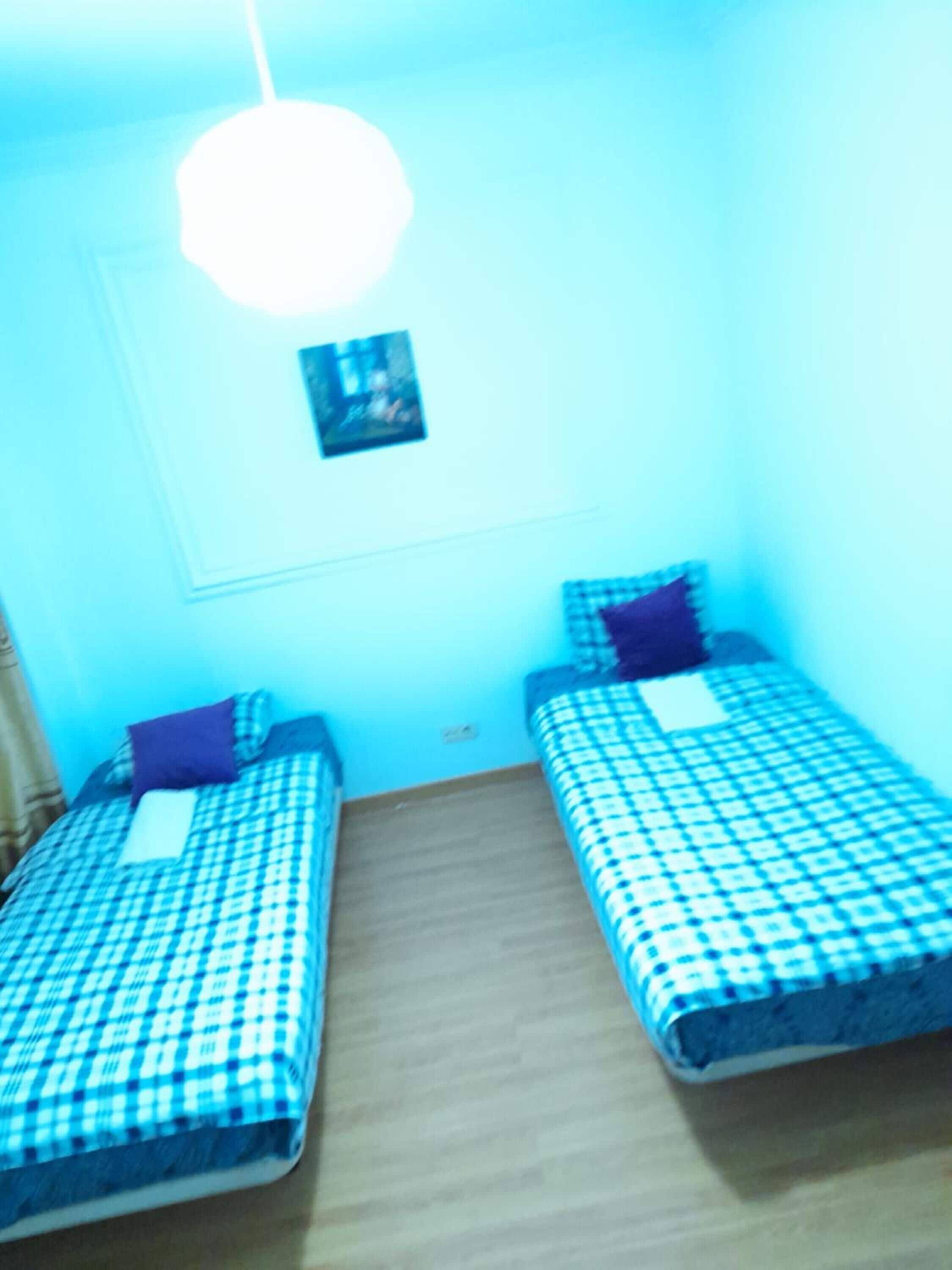 Une petite pièce avec deux lits, chacun recouvert d’une couverture à carreaux et agrémenté d’un oreiller. Un seul luminaire est fixé au plafond, et un petit tableau est visible sur le mur.