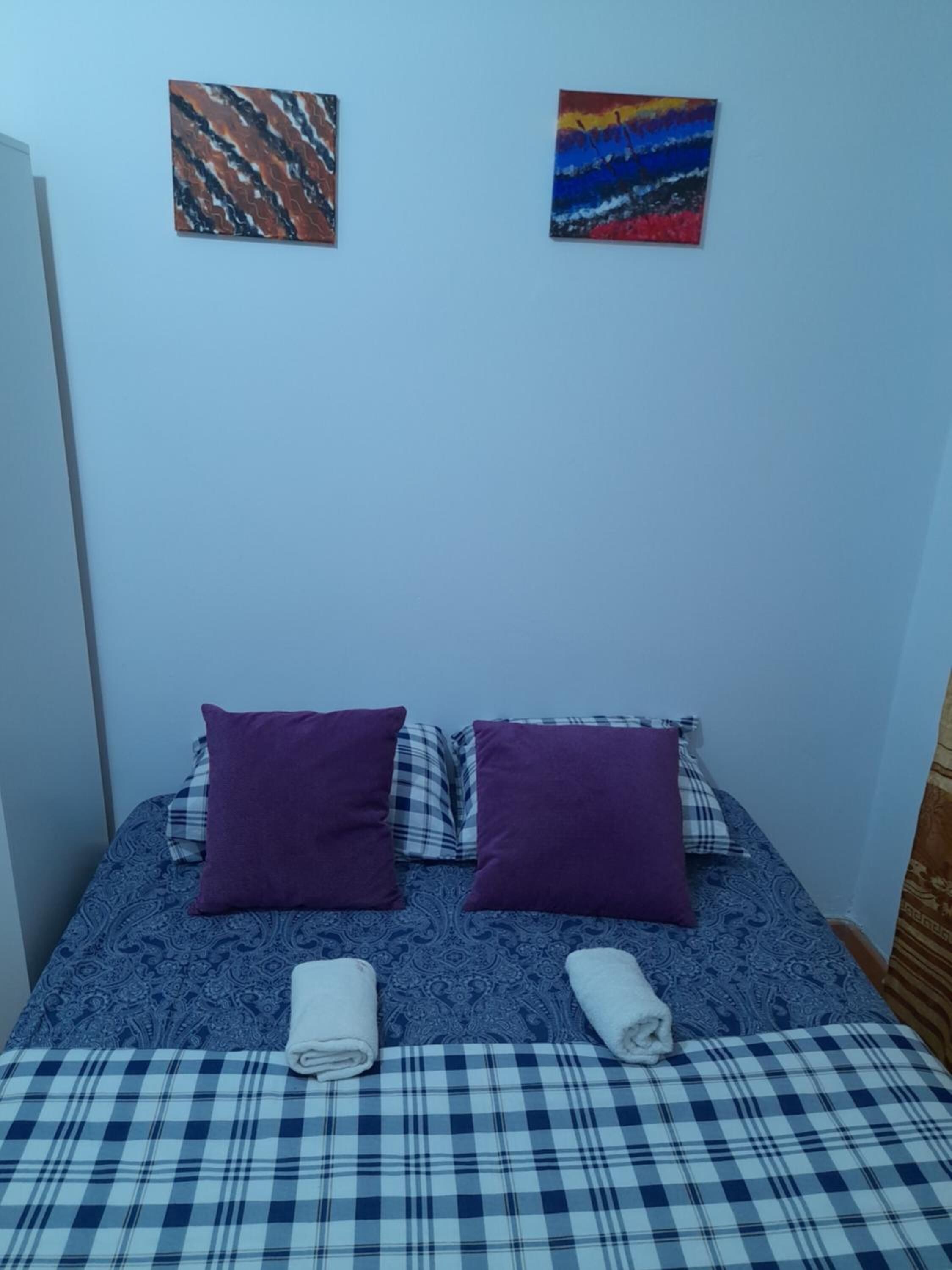 Una cama con una colcha azul y blanca a cuadros, dos almohadas moradas y dos toallas enrolladas. Encima de la cama, hay dos cuadros en la pared.