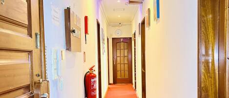 Een hotelgangen met een rood tapijt, houten deuren en een brandblusser aan de muur.