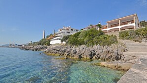 Una propiedad costera con aguas azules cristalinas, costa rocosa y una escalera que lleva a un edificio con balcones.
