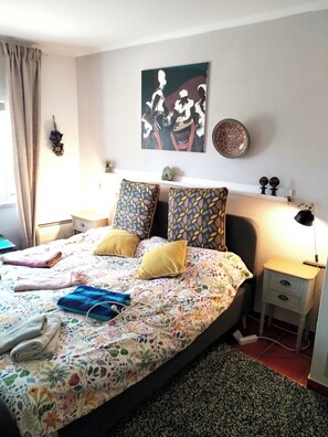 Ein Schlafzimmer mit floralem Bettbezug, zwei gemusterten Kissen, einem Bild an der Wand, einem Nachttisch mit Lampe und einer kleinen Ablage mit Dekorationen.