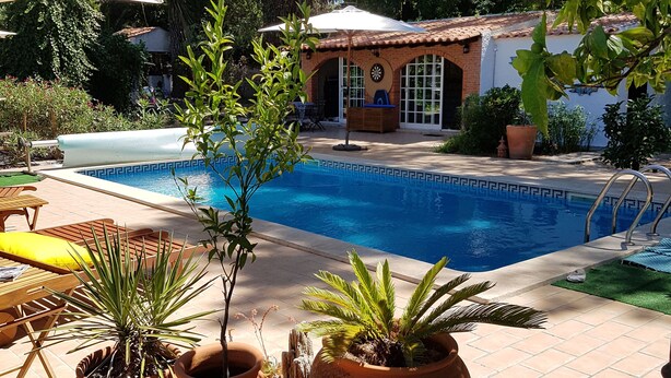 Un espace piscine agrémenté de jardinières en terre cuite, une terrasse avec une table et des chaises, et un bâtiment doté d’un toit en tuiles.