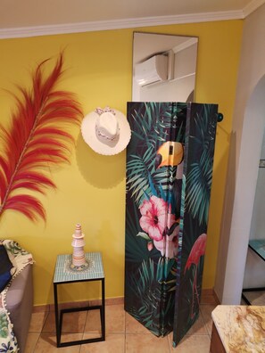 Quarto com parede amarela, uma pena decorativa, um chapéu, uma mesa pequena com tampo estampado e uma porta com estampa tropical.