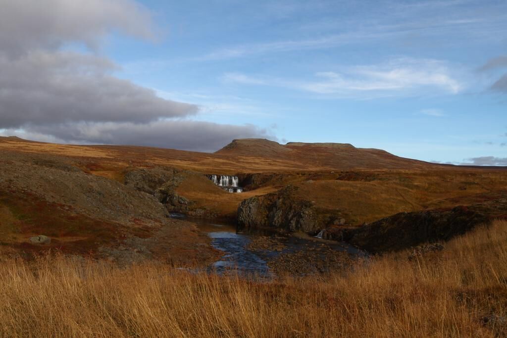 Un paisaje natural y aislado con un pequeño edificio, posiblemente una cabaña, rodeado de colinas y un arroyo.