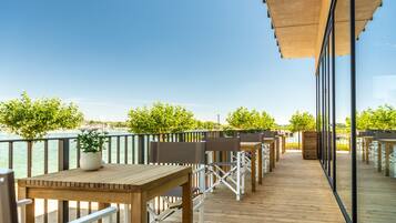 Une terrasse en bois, agrémentée de tables et de chaises, qui offre une vue sur une étendue d’eau et sur la verdure.