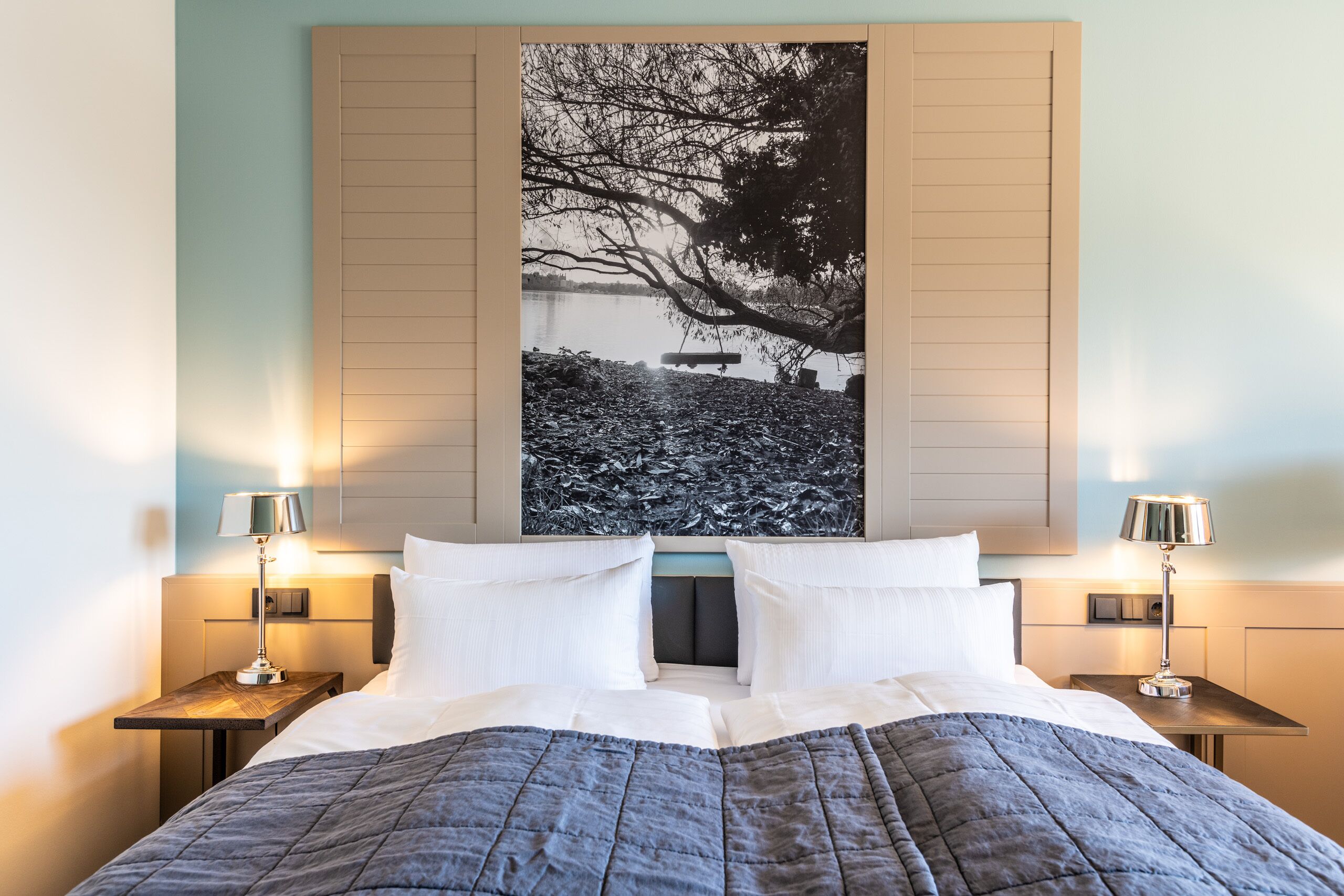 Ein ordentlich bezogenes Bett mit einer dunklen Steude, zwei Nachttische mit Lampen und ein großes Schwarz-Weiß-Foto an der Wand.