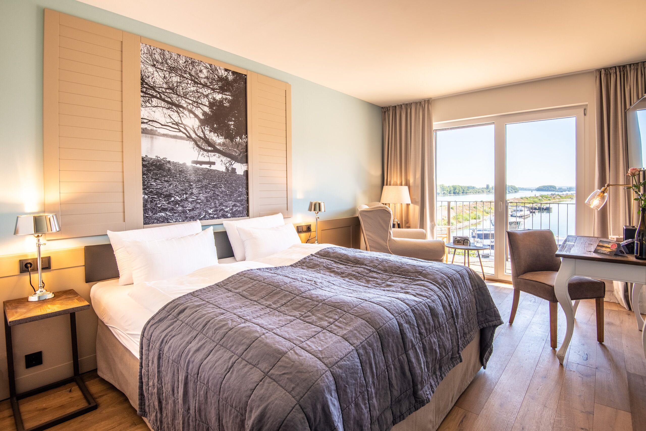 A hotel room with a large bed, a desk with a chair, a view of a body of water, and a painting on the wall.