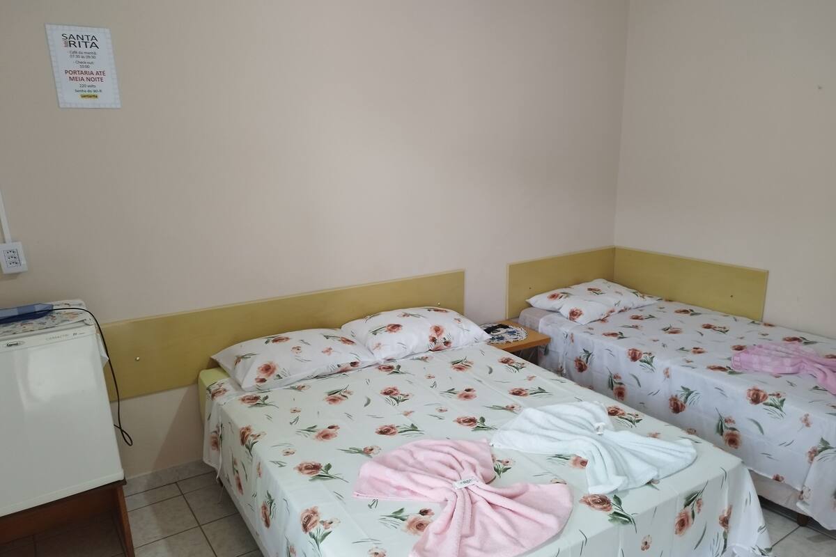 A hotel room with two beds, each with floral bedding and a pink towel. There is a small table with a phone and a sign on the wall.