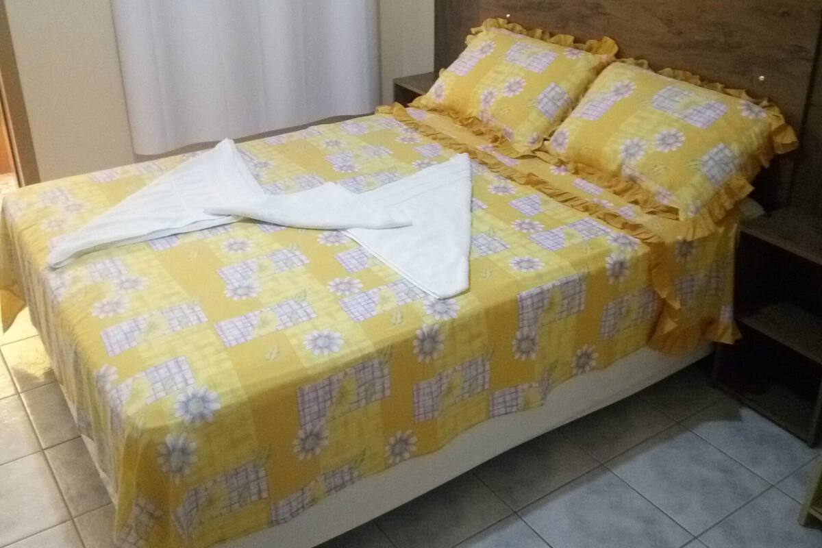 A bed with a yellow floral bedspread and two pillows, a wooden headboard, and a white curtain.