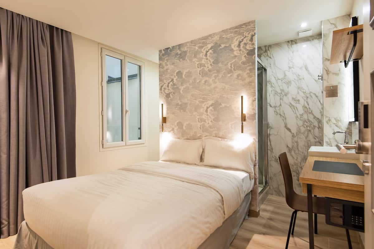 A hotel room with a bed, a desk with a chair, a bathroom with a mirror, and a window with curtains.