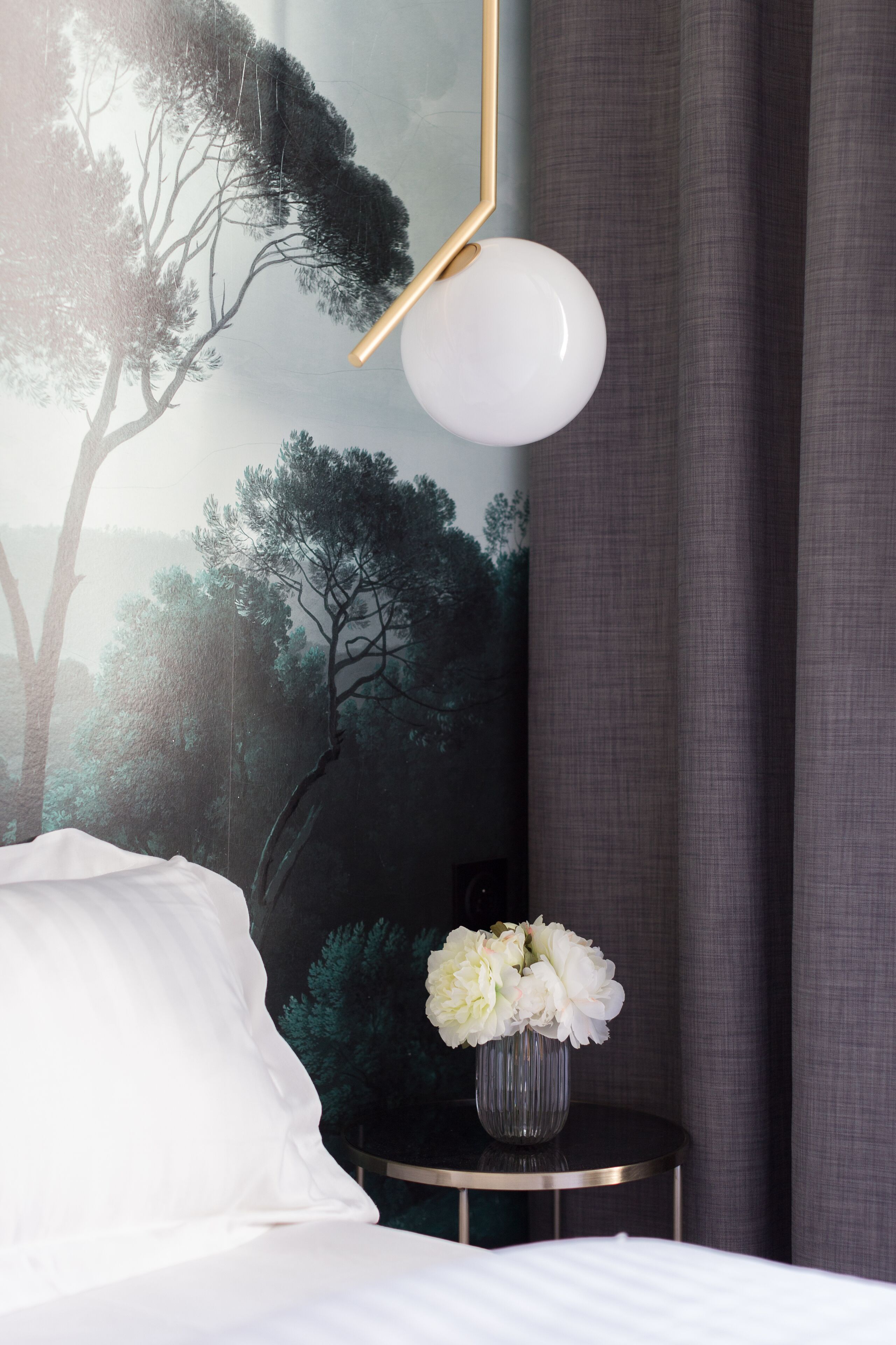 Quarto com cama, um vaso de flores, luminária na parede e pintura de árvores.
