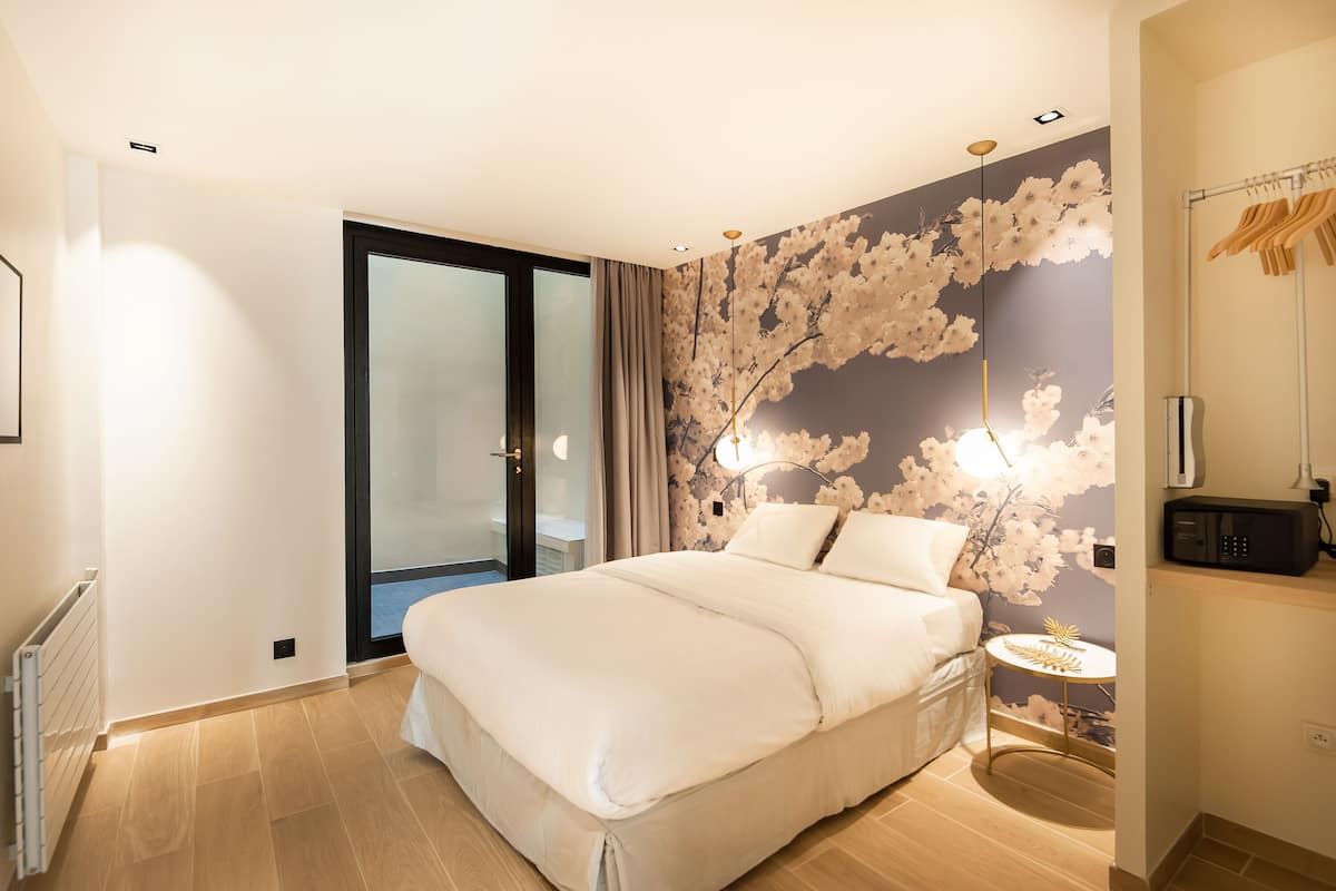 A modern bedroom with a large bed, a glass door leading to a balcony, a wall with floral wallpaper, and a small round table with a lamp.