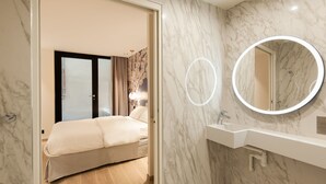 Una habitación de hotel moderna con baño de mármol, cama con ropa de cama color beige y un espejo redondo con iluminación alrededor.