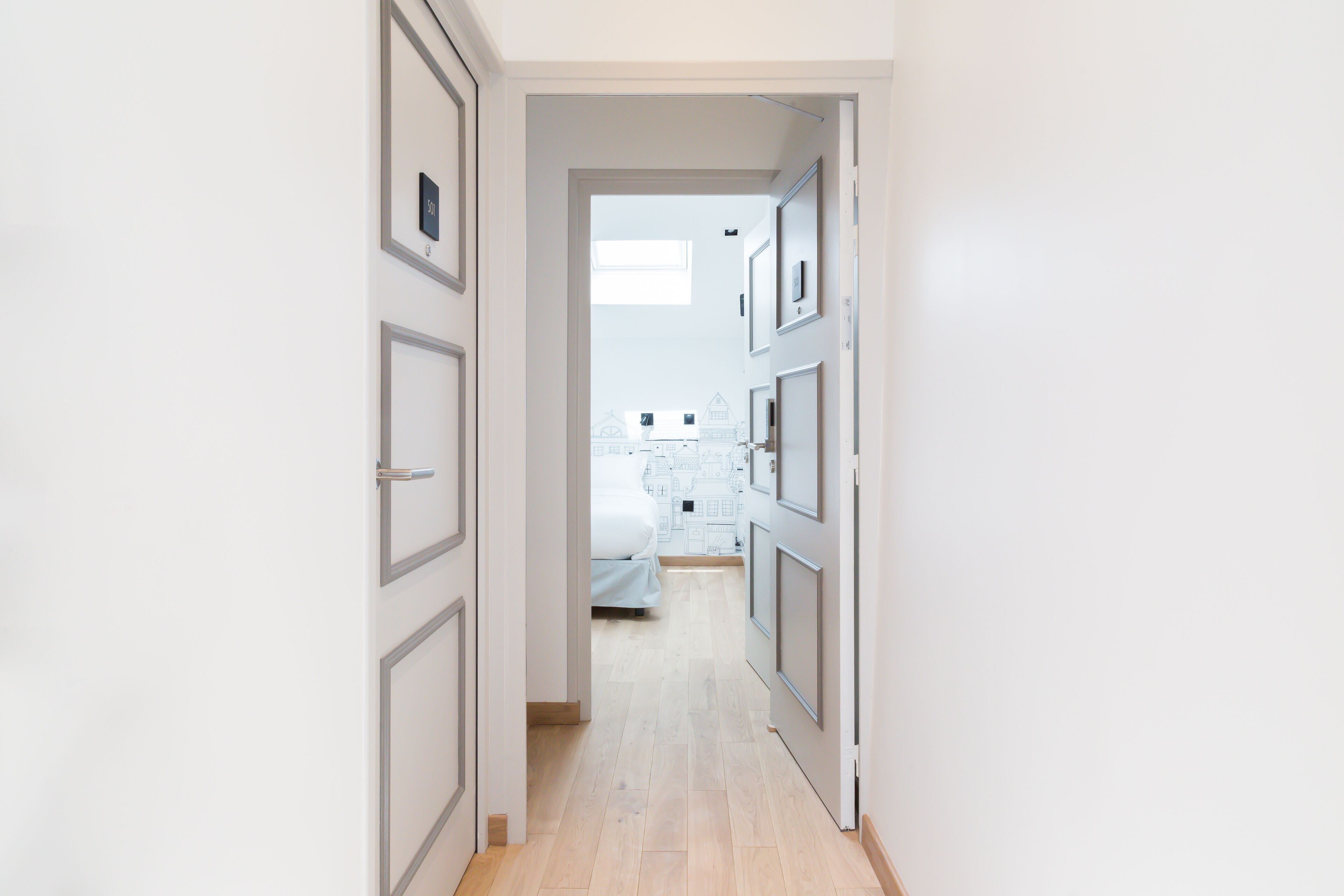 A narrow hallway with light wooden flooring, leading to a bedroom with a bed and a window.