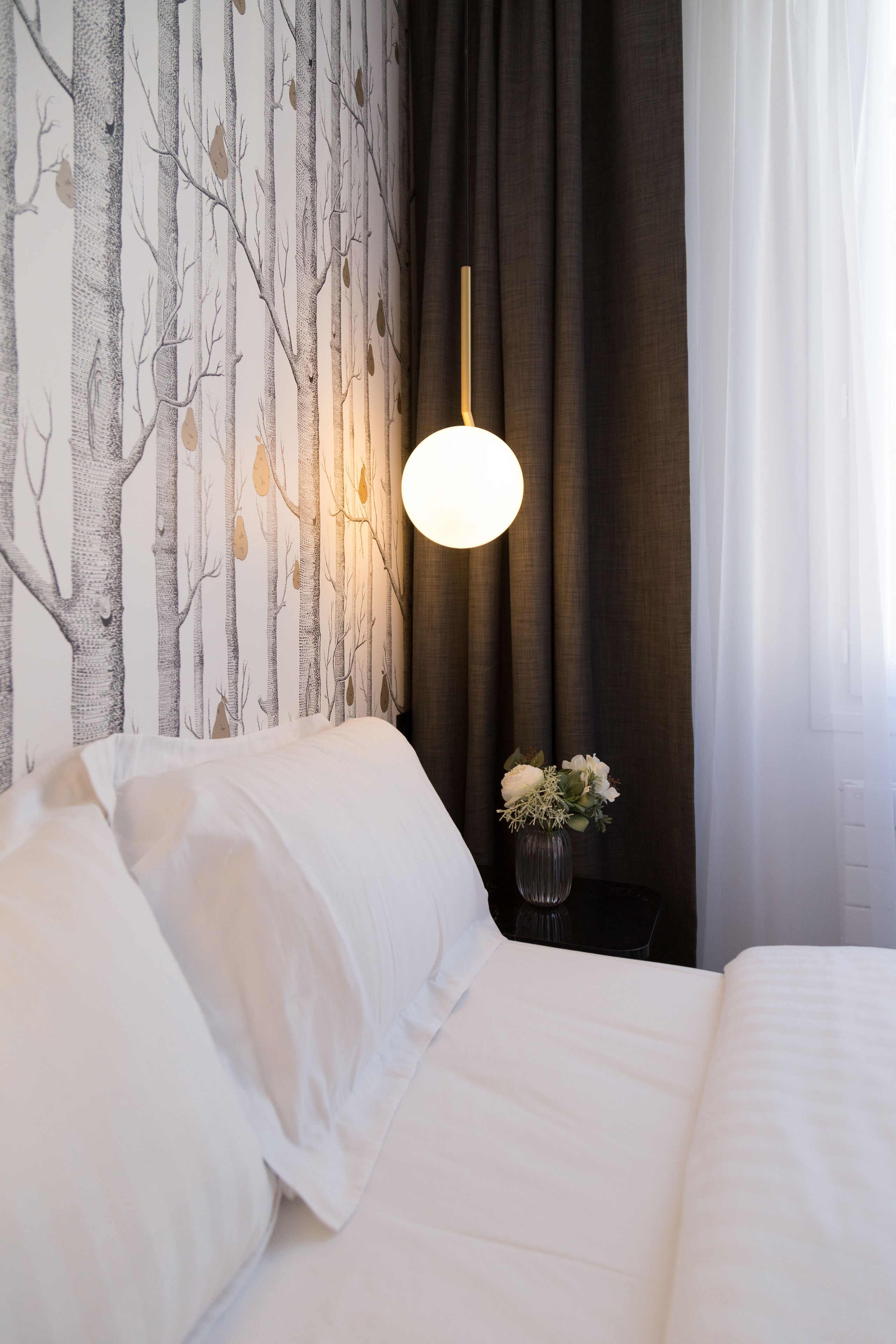 A neatly made bed with white linens, a wall with a tree pattern wallpaper, a hanging light fixture, and a vase with flowers on a bedside table.