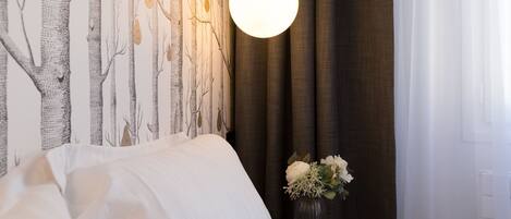 A neatly made bed with white linens, a wall with a tree pattern wallpaper, a hanging light fixture, and a vase with flowers on a bedside table.