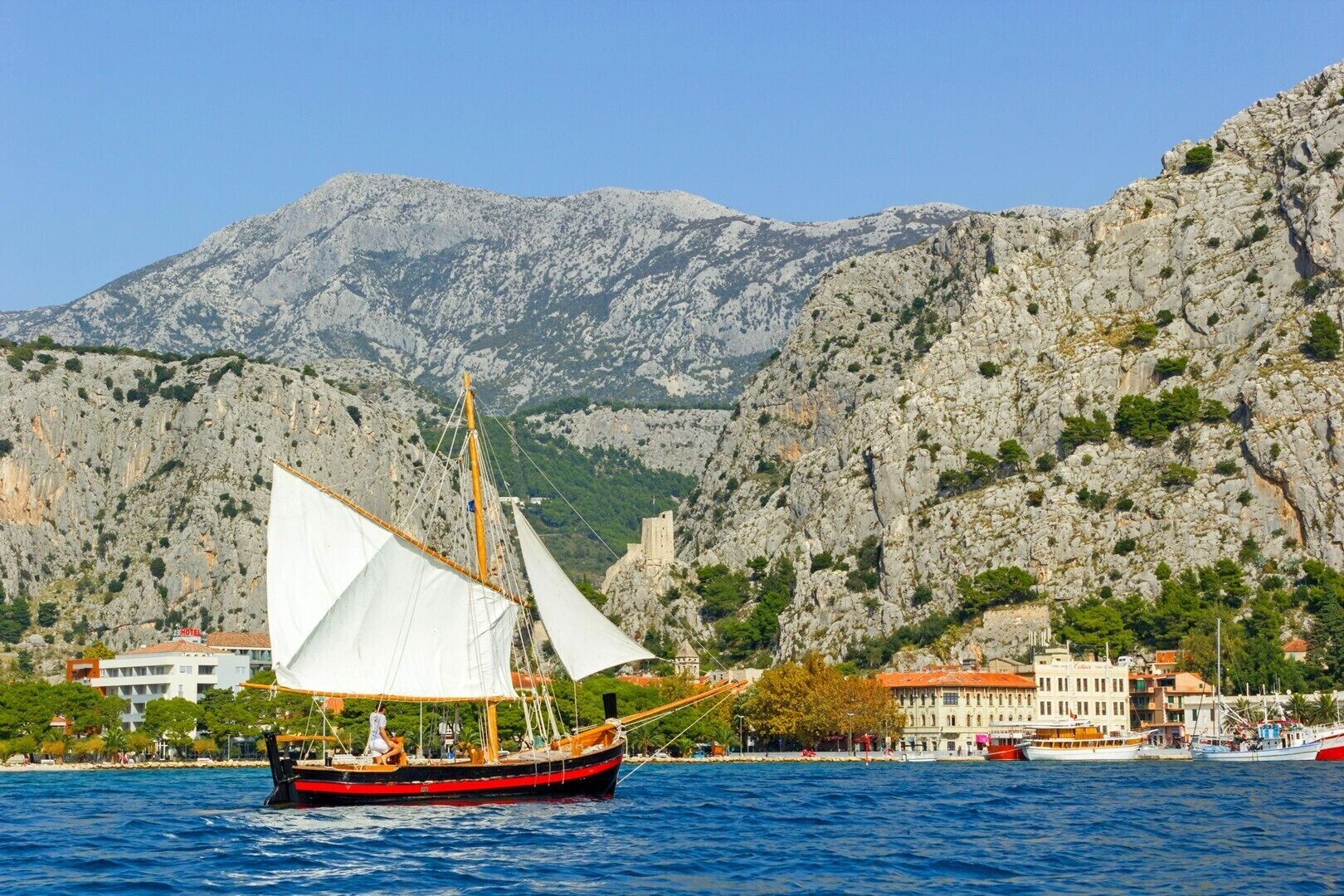 A traditional sailing boat with white sails, docked near a coastal town with rocky mountains in the background.