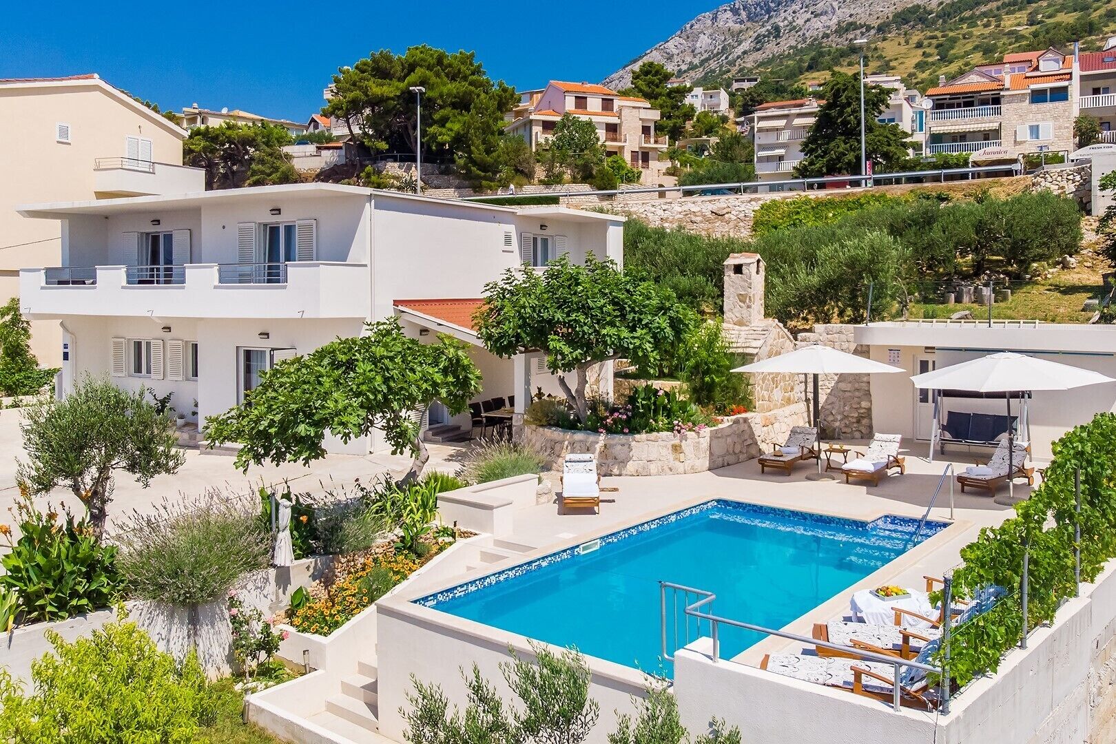 A swimming pool with lounge chairs and umbrellas, surrounded by greenery and buildings.