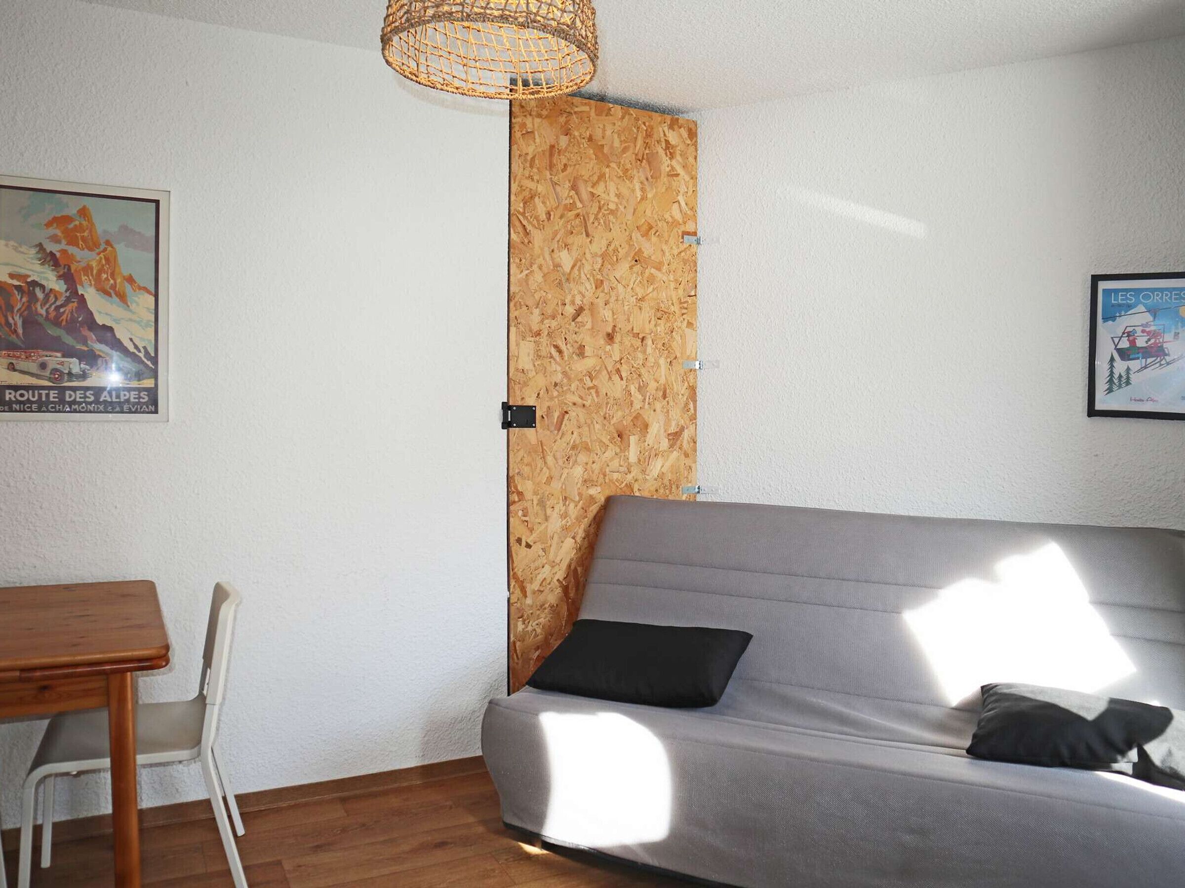 A modern living space with a grey sofa, a wooden wall panel, a dining table with chairs, and two framed pictures on the walls.