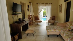 Ein Wohnzimmer mit Fernseher, einer Couch, Sesseln und Blick auf ein Schwimmbecken.