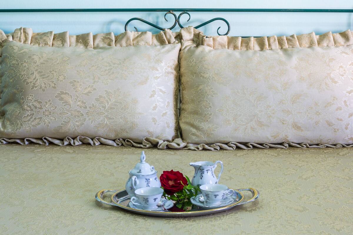 A bed with a decorative headboard, a set of white and gold embroidered pillows, and a tray with tea set and a red rose.