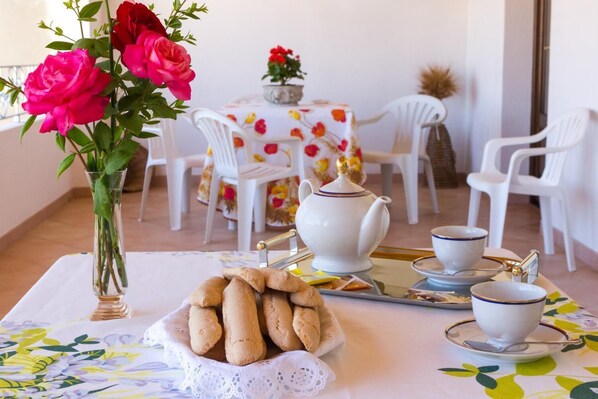 A table set with a teapot, cups, and a bowl of cookies, with a vase of flowers and a floral tablecloth. - B&B Casa Titti (Cabras)