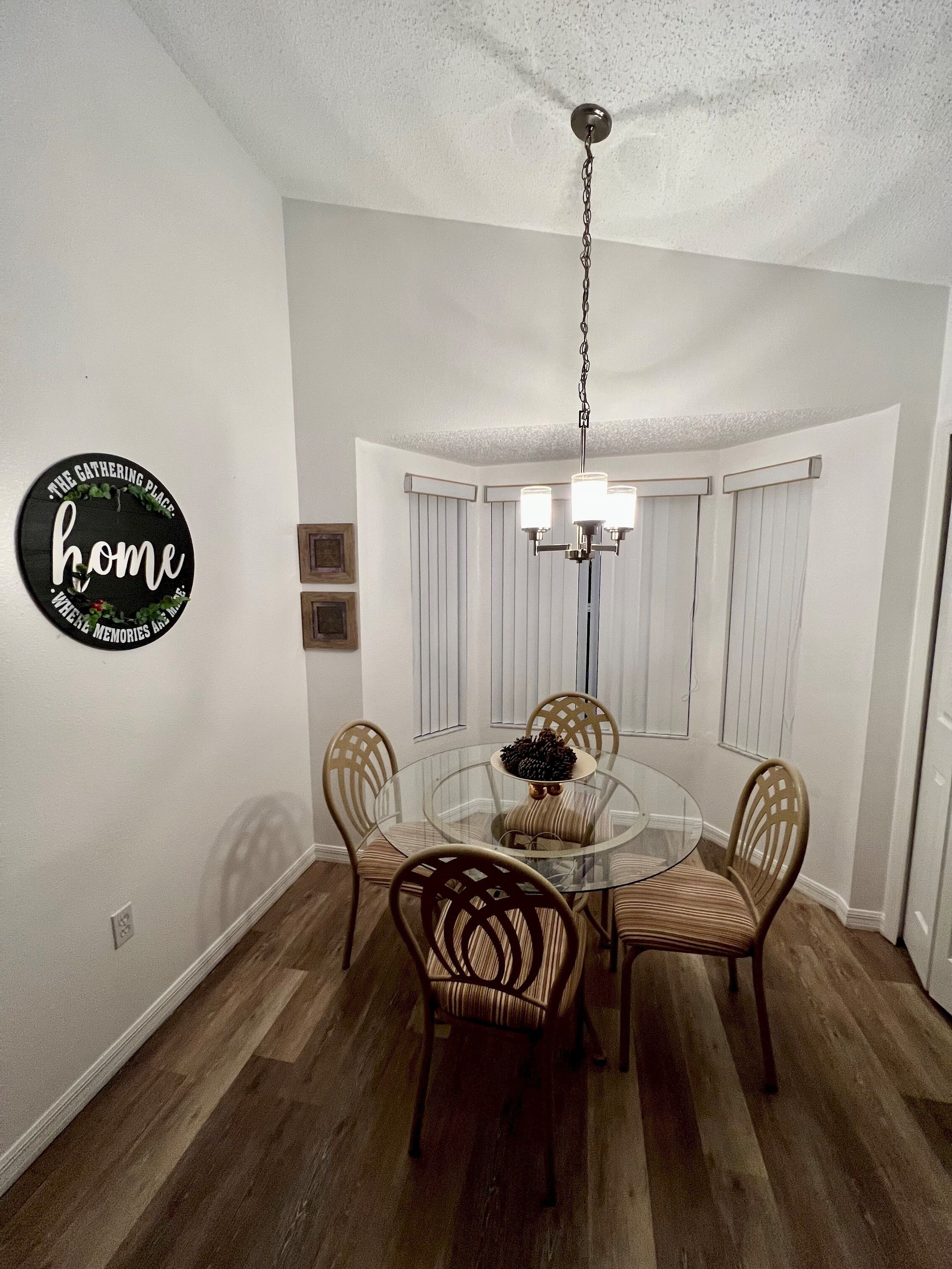 A dining area with a round glass table, four wicker chairs, a chandelier, and a wall sign that reads "The Gathering Place Home."