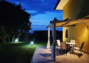 An outdoor patio area with a table and chairs, a pergola, and a well-lit garden.
