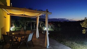 Un patio al aire libre con mesa y sillas, una pérgola con luces y vistas del paisaje.