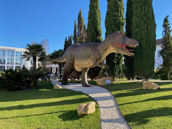 Une statue de dinosaure dans un jardin aménagé, avec un sentier, de grands arbres et un bâtiment en arrière-plan.